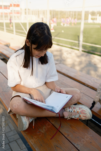 Obraz na płótnie A young girl sits cross-legged on wooden benches, engrossed in reading a book. In the background, kids play on a sports field, highlighting a scene of education and leisure. w salonie