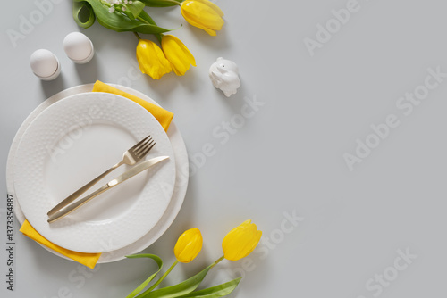 Easter table setting with yellow tulips, eggs , white plate, bunny on light gray background. Spring Easter celebration. View from above. Copy space.