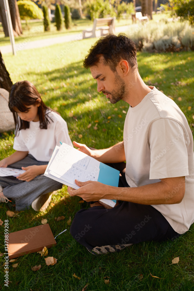A father and daughter spend quality time studying in a sunny park. They are seated on the grass, reviewing papers, enjoying a peaceful and educational moment together amid nature's beauty.