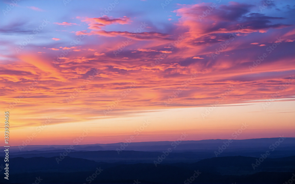 Obraz premium A vibrant sky ablaze with colors at dusk or dawn, showcasing hues of pink, orange, and blue. Clouds are painted with light above a dark silhouette of distant mountains at the horizon.