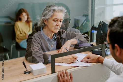 Foto Elderly woman signing official documents at banking counter with teller assistin