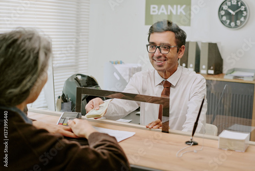 Bank teller wearing glasses and a white shirt smiling while assisting customer by handing currency over counter in office