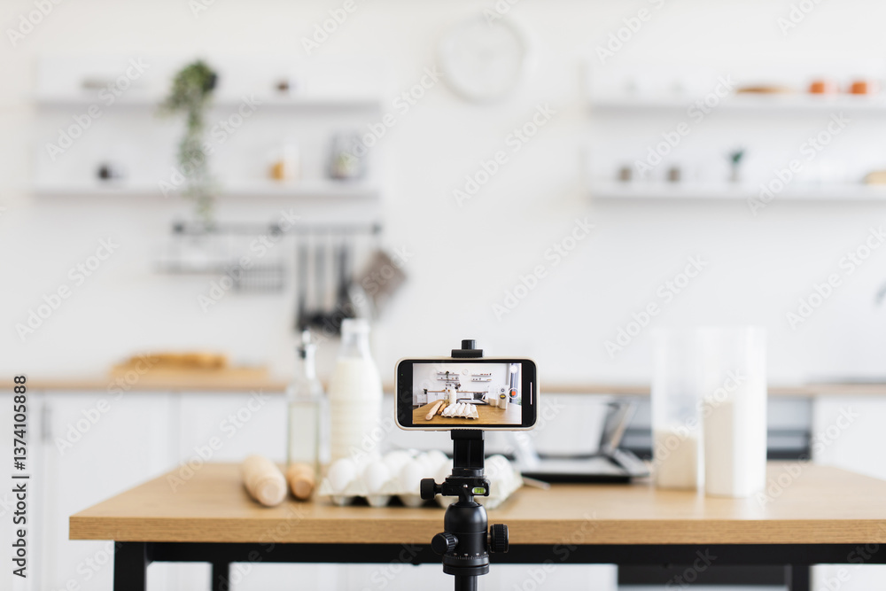 Bright kitchen scene showing a phone on a tripod recording a baking video. Ingredients include eggs, milk, flour, and other supplies, suggesting culinary content creation with modern camera equipment. Bright kitchen scene showing a phone on a tripod recording a baking video. Ingredients include eggs, milk, flour, and other supplies, suggesting culinary content creation with modern camera equipment.