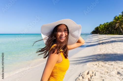 Beautiful happy lady wearing straw hat at beach with turquoise ocean on background, looking at camera and laughing, enjoying tropical vacation