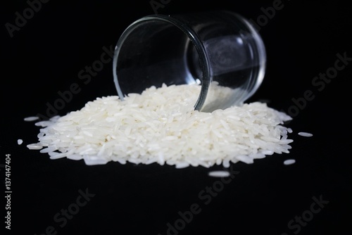 A small, clear glass is tilted to the side against a pitch-black background. The cup is filled with a pile of raw, white rice, which is piled up in front of the cup. The rice grains are long and shiny