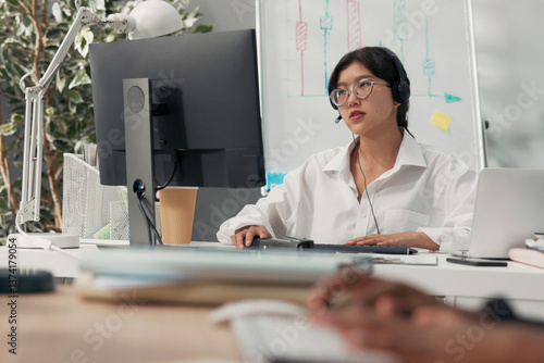 Beautiful elegant woman in glasses with Asian Korean beauty sits in office of company with headphones microphone on ears in front of computer conducts client consultations insurance explains