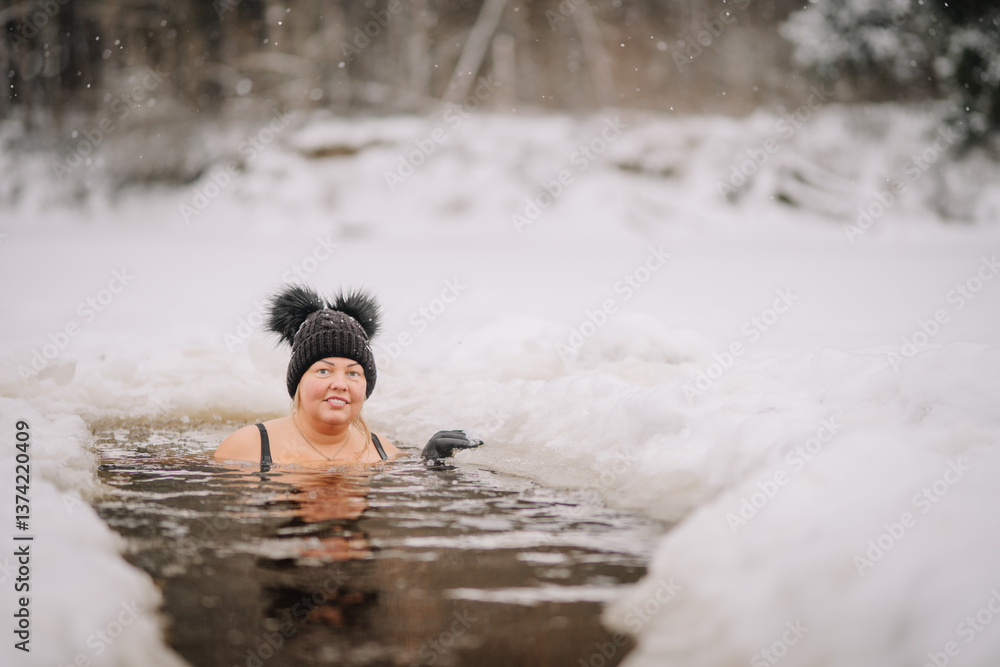 Fototapeta premium Smiling woman in black swimwear and winter hat enjoys an ice bath in a snowy outdoor setting, surrounded by frozen landscape and falling snowflakes..