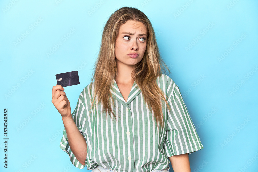 Young blonde with a credit card in blue studio confused, feels doubtful and unsure. Young blonde with a credit card in blue studio confused, feels doubtful and unsure.