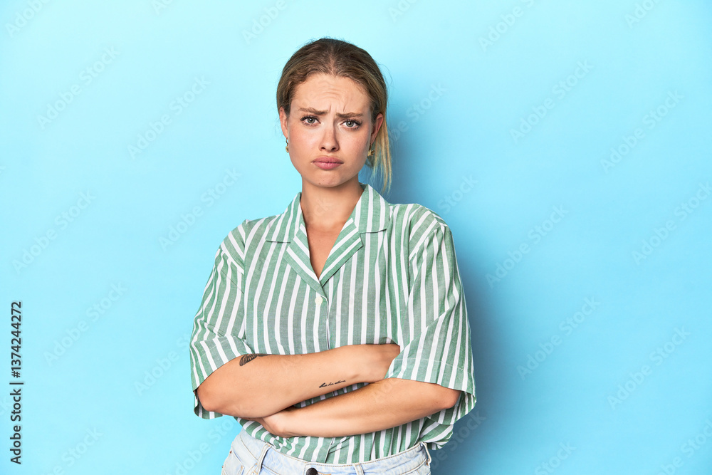 Blonde young woman in green striped shirt on blue background frowning face in displeasure, keeps arms folded. Blonde young woman in green striped shirt on blue background frowning face in displeasure, keeps arms folded.