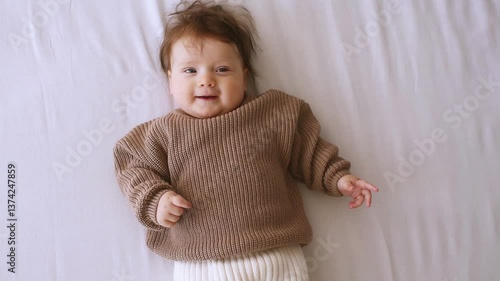Smiling baby girl wearing warm knitted sweater and pant lying in bed over white blanket. Babyhood. 