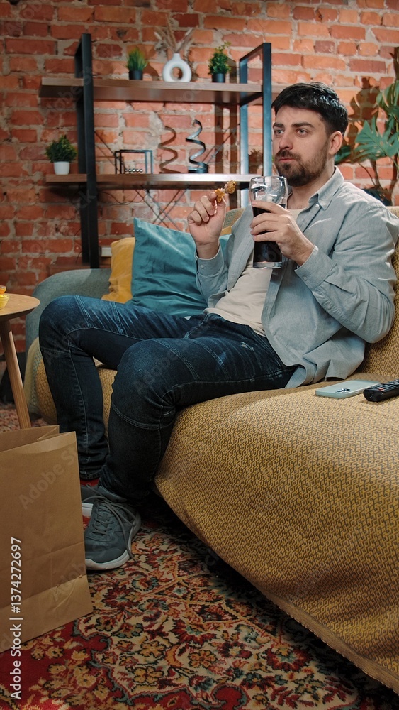 Fototapeta premium Man spending time at home with fast food delivery, sits on couch watching tv, eating dipping fried chicken in sauce, drinking cola, enjoying the taste.