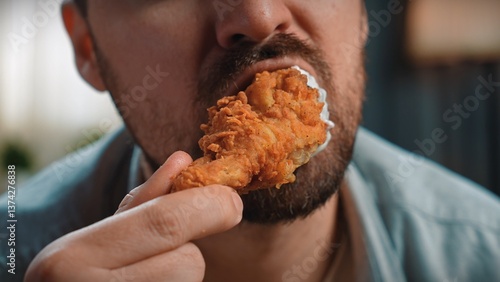 Close up shot of bearded man face man holding and biting chewing fried chicken leg, enjoying eating fast food.