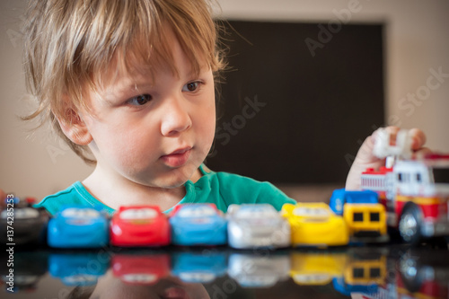 A little boy carefully lines up colorful toy cars and a fire truck on a table. His focused expression shows he is deeply engaged in imaginative play in a modern room