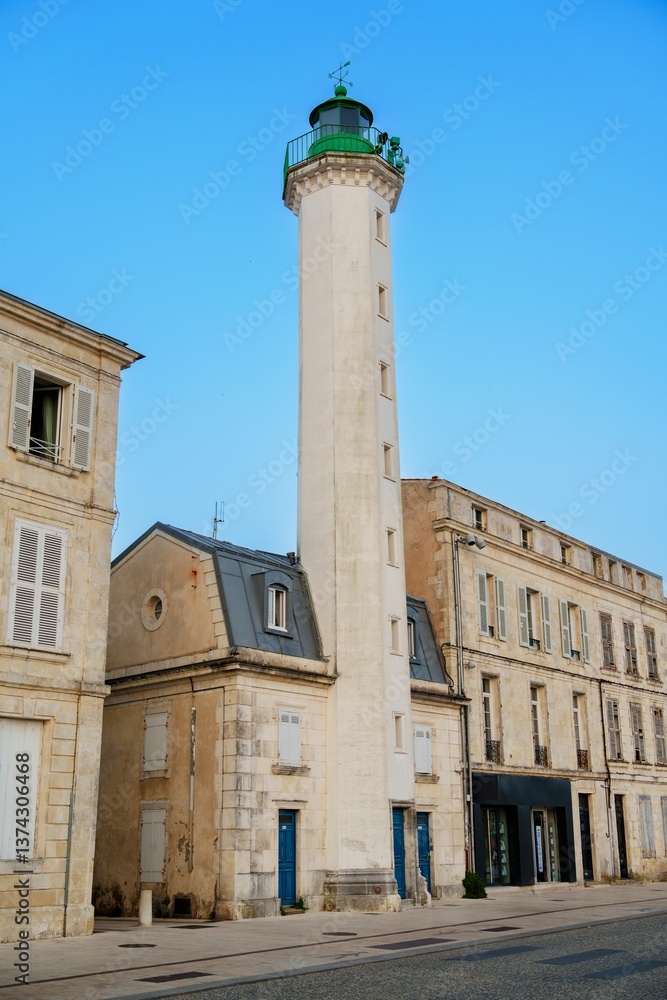 Fototapeta premium An old lighthouse proudly stands near historic harbor buildings, surrounded by calm waters and under a clear blue sky, creating a picturesque scene that embodies maritime history and charm.
