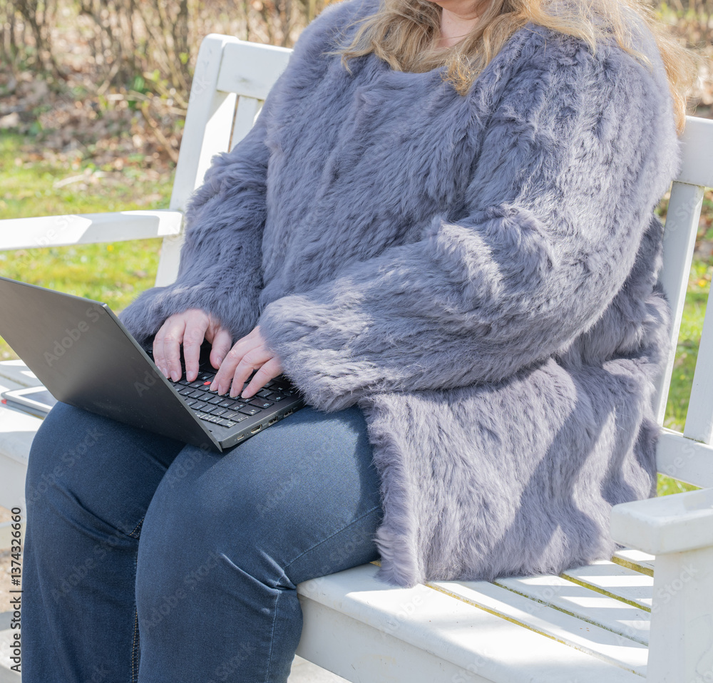 Frau in einer Kaninchenfell Jacke arbeitet im Garten mit einem Notebook im Homeoffice