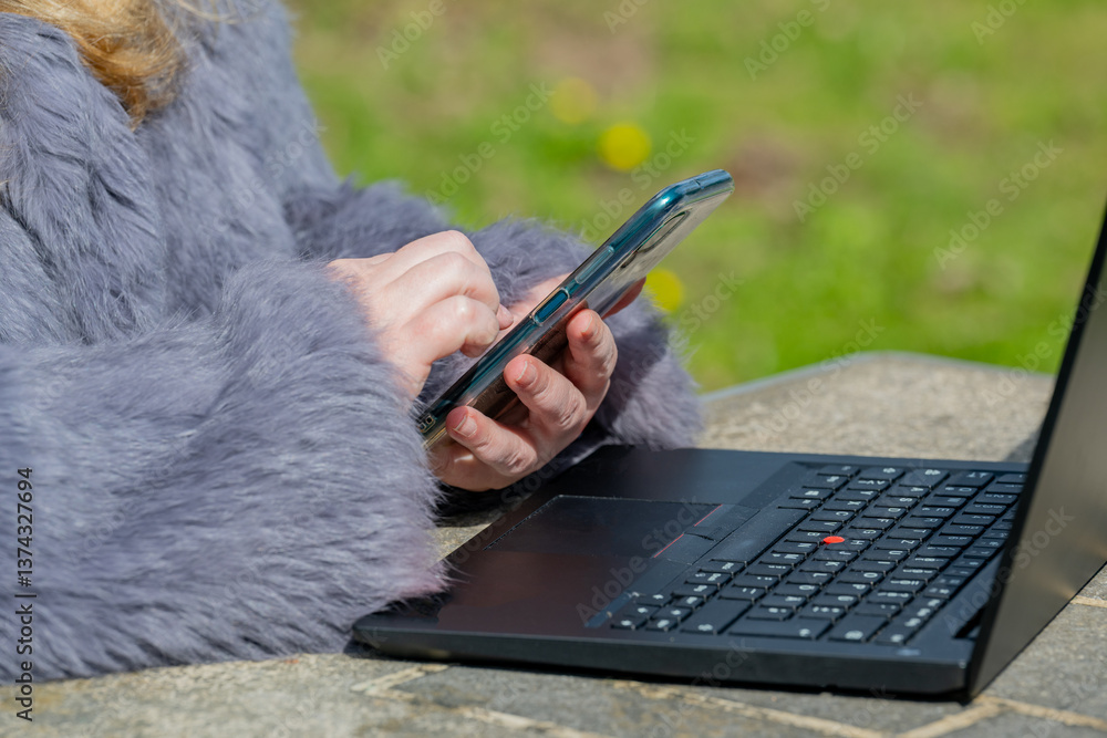 Frau in einer Kaninchenfell Jacke arbeitet im Garten mit einem Notebook und Handy im Homeoffice