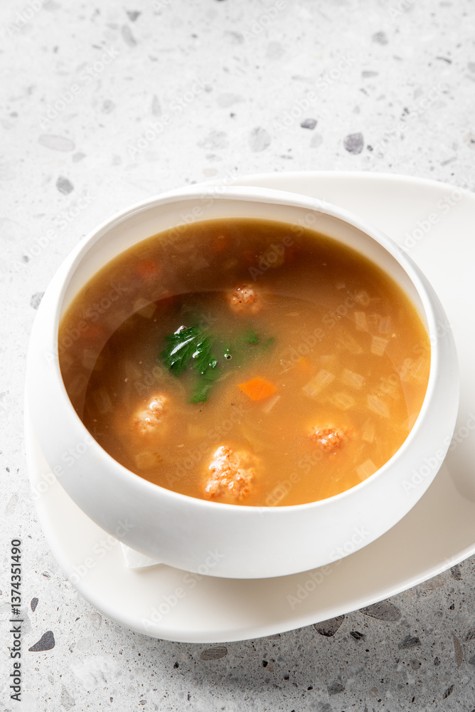 A bowl of clear soup with meatballs, diced carrots, onions, and green vegetables on a white plate, placed on a speckled gray countertop, perfect for culinary presentations. A bowl of clear soup with meatballs, diced carrots, onions, and green vegetables on a white plate, placed on a speckled gray countertop, perfect for culinary presentations.