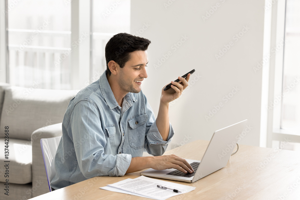 Smiling man speaking on speakerphone seated at desk with laptop, using AI virtual assistant app, browse internet, searching information, enjoy easy, convenient usage of modern wireless technologies