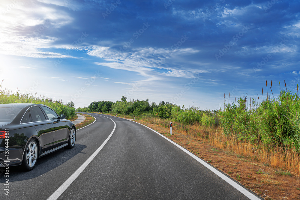 A black sedan is parked on the side of a curved, tree-lined highway under a bright blue sky with scattered clouds, highlighting a serene moment in a scenic countryside journey.