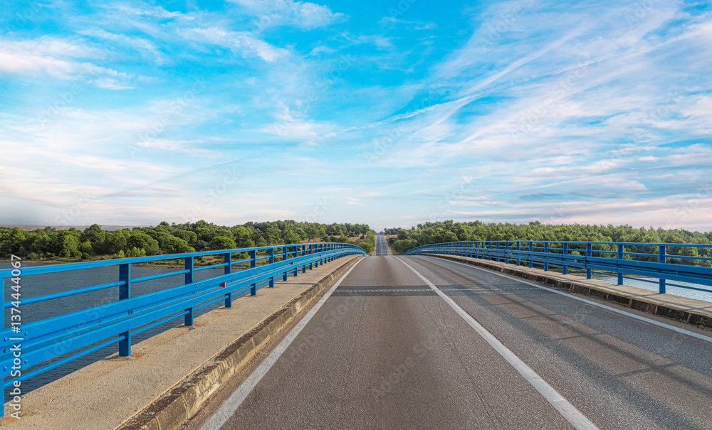 A long, straight bridge stretches over a body of water under a bright blue sky, creating a scenic and peaceful road trip atmosphere.