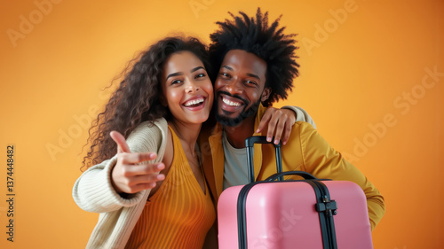 Smiling young couple embracing in front of a pink suitcase, posing with enthusiasm and joy against an orange background. Happy couple with luggage ready to travel against a colorful background.