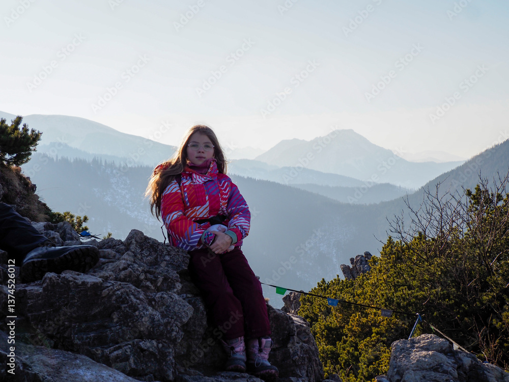 Naklejka premium Young Girl in Pink Winter Jacket Sitting on Mountain Rock with Panoramic Mountain View