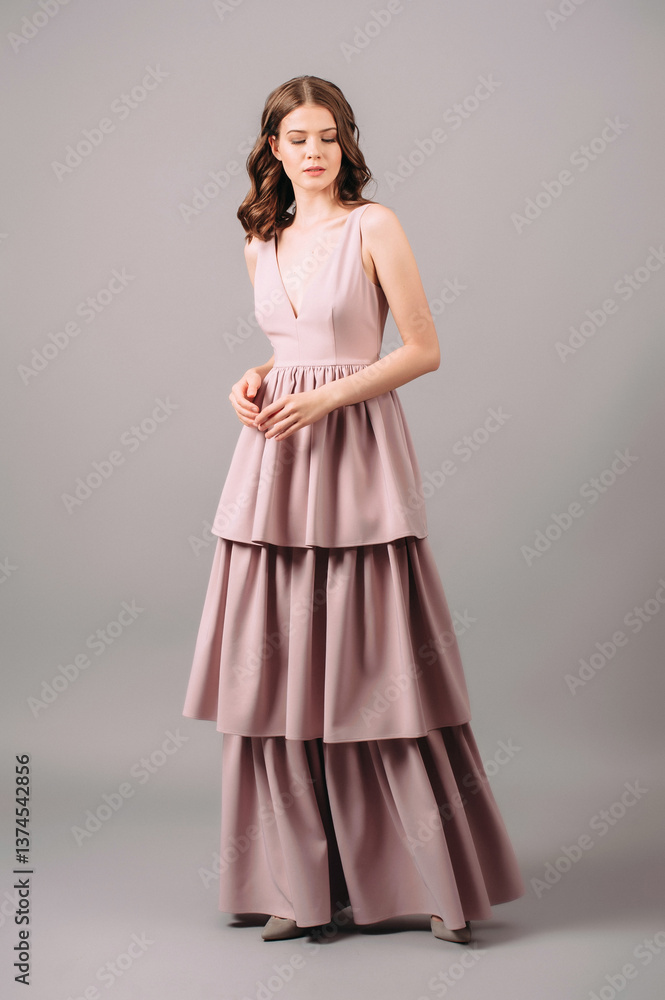 Fototapeta premium Romantic portrait of brunette woman in powder evening dress walking in studio. Bridesmaid's fashion. Dust pink plunge neckline sleeveless maxi gown with tiered skirt.Summer feminine look for hen party