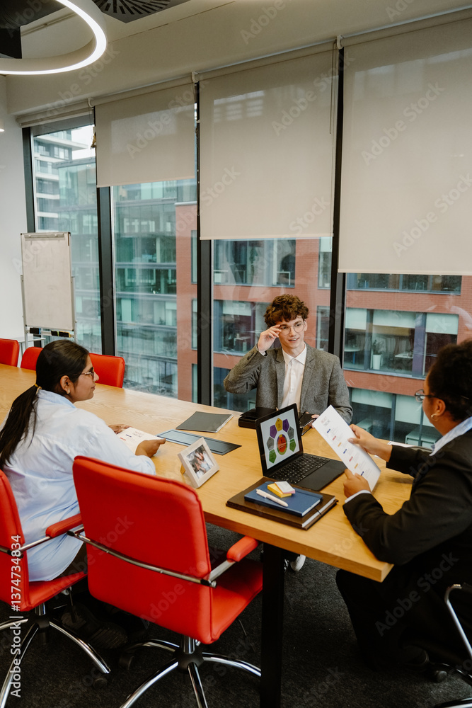 Obraz premium Two female employees sitting at a table and holding papers while looking at a male intern sitting next to them and smiling, in the office, during an internship
