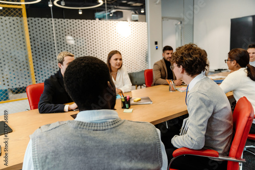 During a meeting in a business room, seven colleagues of different ethnicities are sitting at a long table on red chairs. Concept of negotiations, team discussion of work tasks, performance review.