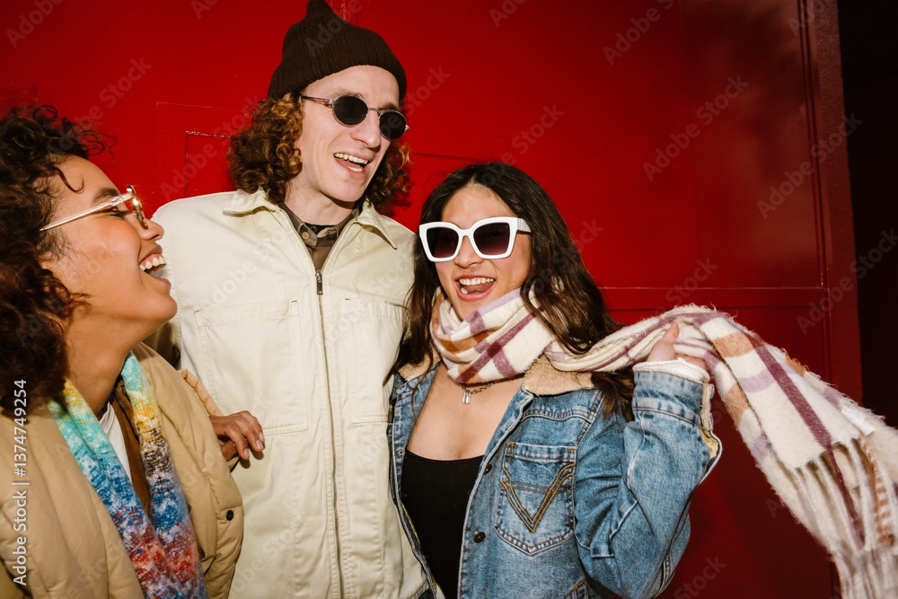 Obraz premium A Black young woman, a White young man, and an Asian young woman laughing and posing together in front of a red wall, enjoying each other's company, wearing trendy jackets and scarves.