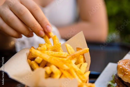 Closeup of young womans hand grabbing a crispy golden fries. Enjoying potato chips with a juicy burger in fast food restaurant on summer day. Selective focus