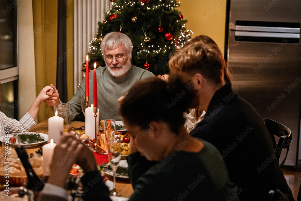 Fototapeta premium A White elder man and his family holding hands during a Christmas dinner, sharing a special holiday moment together around the festive table, with a decorated Christmas tree in the background.