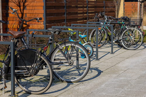 Various bicycles and a small children's bike parked in a metal bike rack on a paved sidewalk near a wooden fence in sunny weather. Urban cycling, eco-friendly transport and outdoor mobility