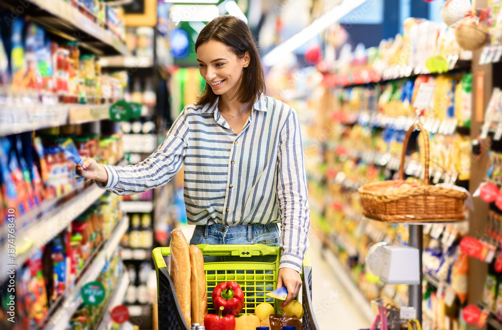 Sale, Shopping, Consumerism And People Concept. Smiling young woman standing with trolley basket full of eco bio products, walking near shelves at grocery store, choosing healthy food in market Sale, Shopping, Consumerism And People Concept. Smiling young woman standing with trolley basket full of eco bio products, walking near shelves at grocery store, choosing healthy food in market