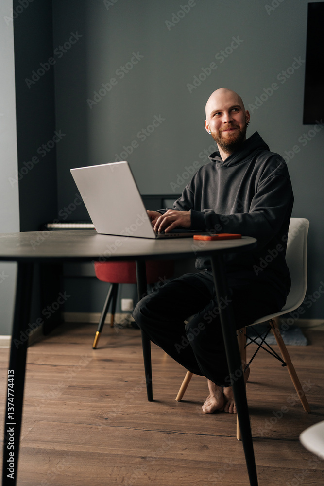 Full length vertical portrait of cheerful programmer male remote working from home office, sitting barefoot at wooden table, typing on laptop computer looking away with happy expression.