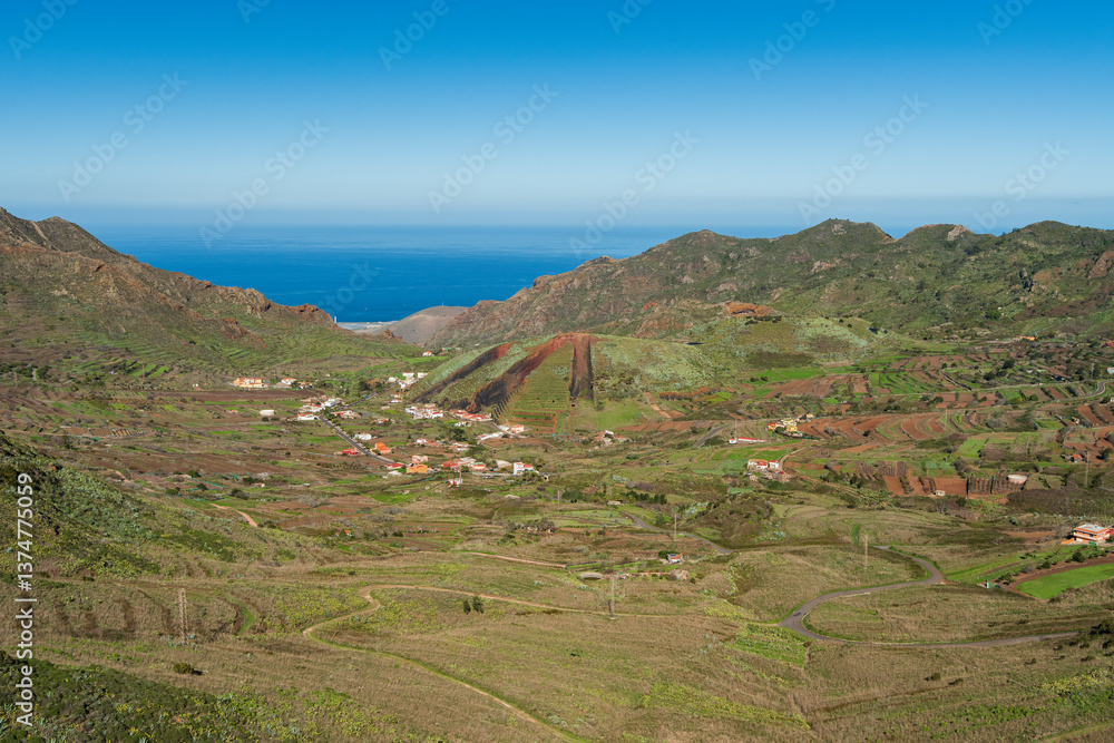 Obraz premium Panoramic view of a rural valley with terraced fields and scattered houses in the mountains of Tenerife, Canary Islands, with the Atlantic Ocean visible in the distance under a clear blue sky