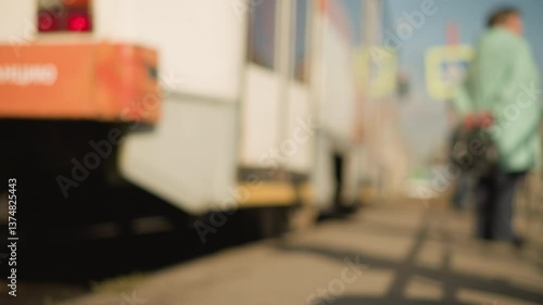 Wallpaper Mural Blurry view of person standing beside tram at tram stop on sunny urban street with soft light casting shadows, tram in motion, pedestrian signs visible in background, evoking calm transit moment Torontodigital.ca