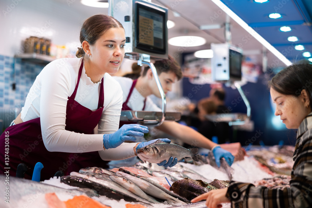 Positive young saleswoman demonstrating seabass to client in fish store with large stock