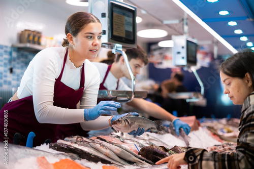 Obraz na płótnie Positive young saleswoman demonstrating seabass to client in fish store with large stock w salonie