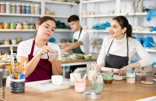 Joyous young women in pinafores sculpting handmade jars of potter-earth material in art studio