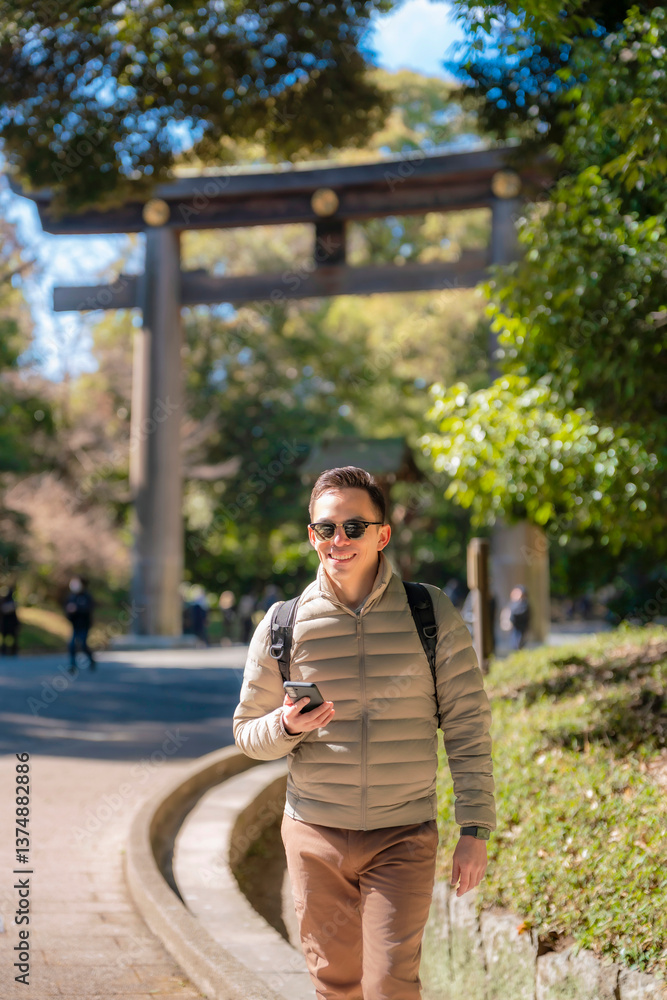 Fototapeta premium Latin young man exploring Meiji Shrine in Tokyo, wearing beige jacket, brown pants, sunglasses, and carrying a backpack with a cheerful attitude.