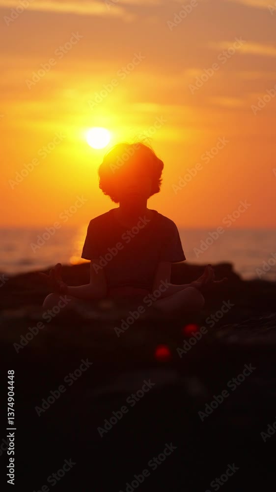 Young Boy In Sukhasana Pose On A Rocky Beach With Orange Sunset Backdrop Young Boy In Sukhasana Pose On A Rocky Beach With Orange Sunset Backdrop