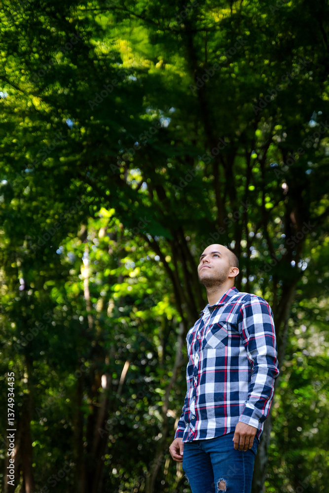 A young man standing in a lush green forest, looking up with a thoughtful expression. He is wearing a plaid shirt and ripped jeans, surrounded by tall trees and vibrant foliage. The sunlight filtering A young man standing in a lush green forest, looking up with a thoughtful expression. He is wearing a plaid shirt and ripped jeans, surrounded by tall trees and vibrant foliage. The sunlight filtering