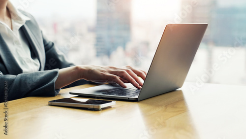 A female business person working with a laptop and smartphone in an office with a city in the background