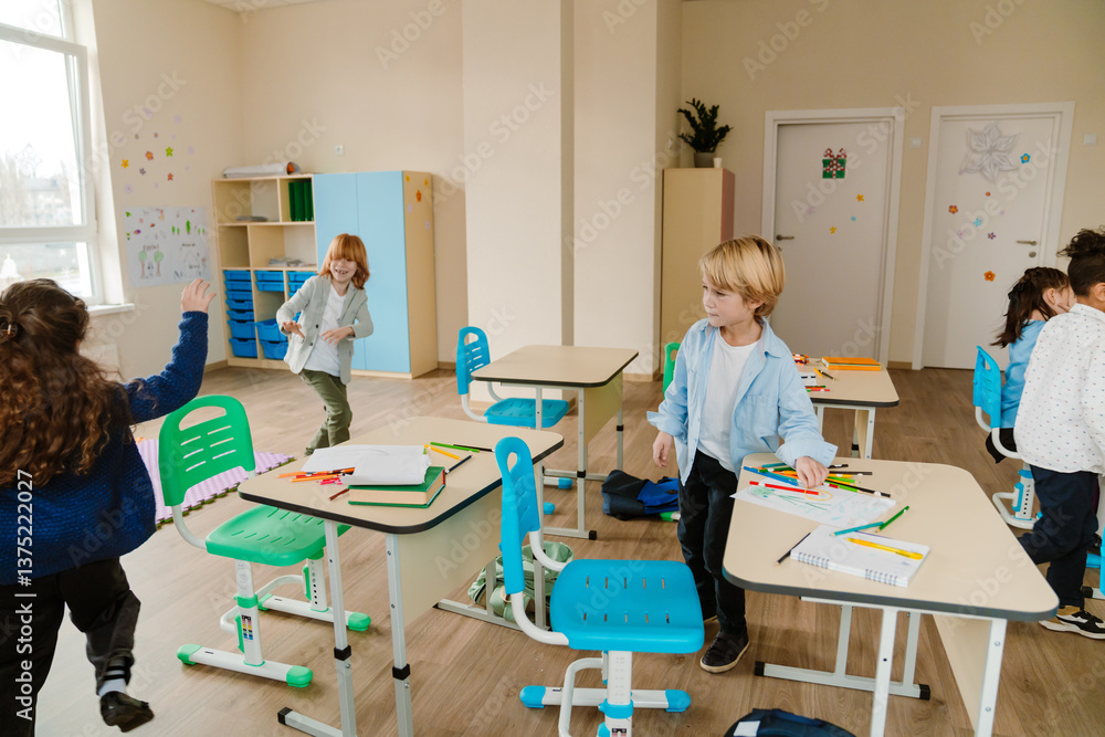 Obraz premium A young, school-aged White boy with blond hair, who is dressed in a button-up shirt, standing next to his desk in a first-grade classroom, as his diverse classmates run around and play on their break