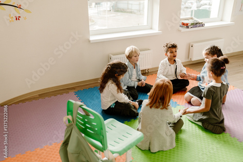 Fotografie A group of diverse elementary school students participates in a talk while sitting in a circle on colorful floor mats in a classroom with white walls and two windows
