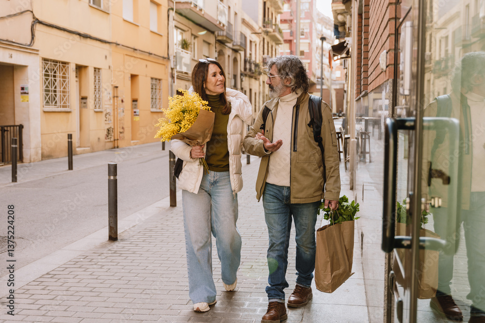 Fototapeta premium A middle-aged White couple of a husband and wife in their 50s, taking a stroll together on a city street, with the woman holding a bouquet of yellow flowers and the man holding a bag of groceries