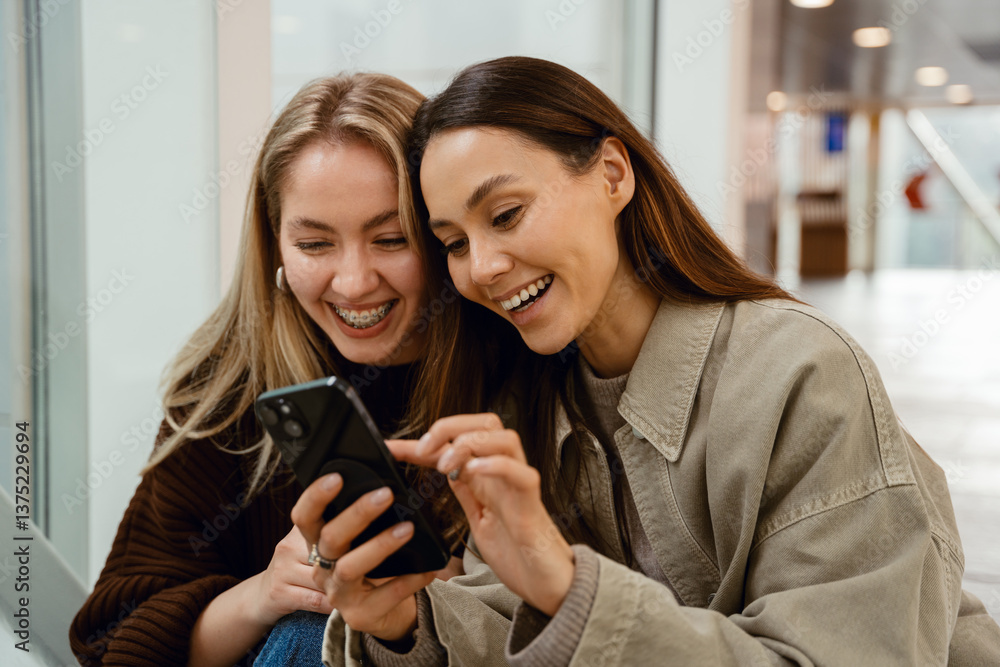 A young brunette White woman in her late 20s, showing something on her phone to her mid-20s, blond White female friend, as they laugh together while sitting indoors, with a window behind them A young brunette White woman in her late 20s, showing something on her phone to her mid-20s, blond White female friend, as they laugh together while sitting indoors, with a window behind them