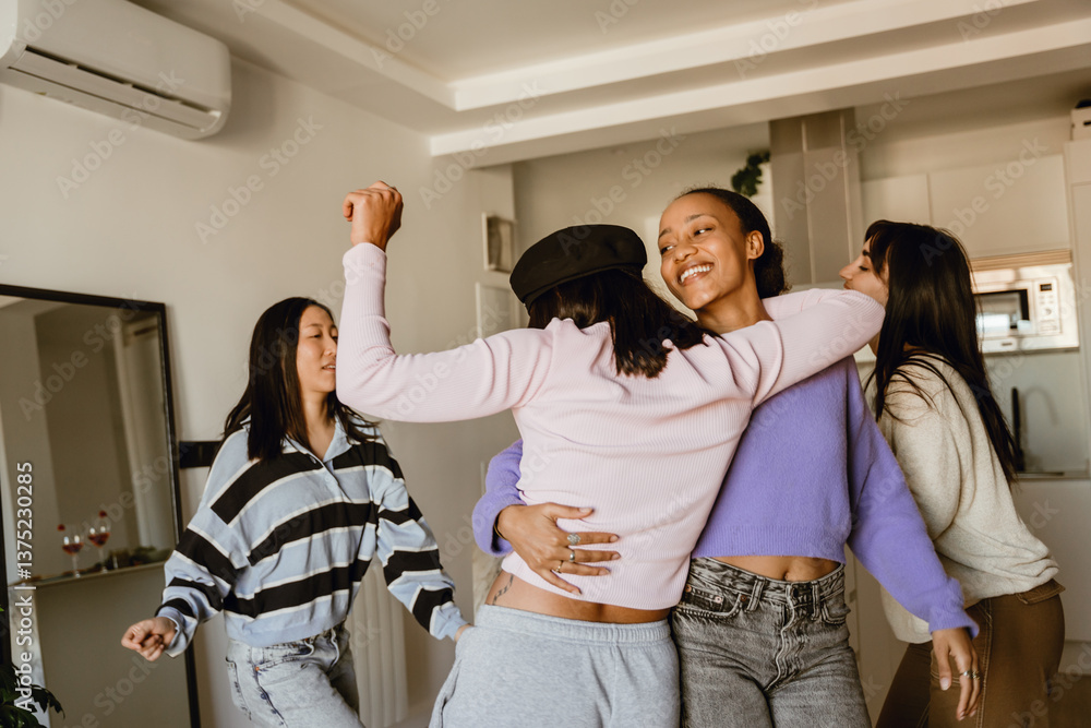 Obraz premium A multiethnic group of four young adult women in casual clothes dancing together in a cozy living room with white walls during a home girls party, two women in the middle hugging each other