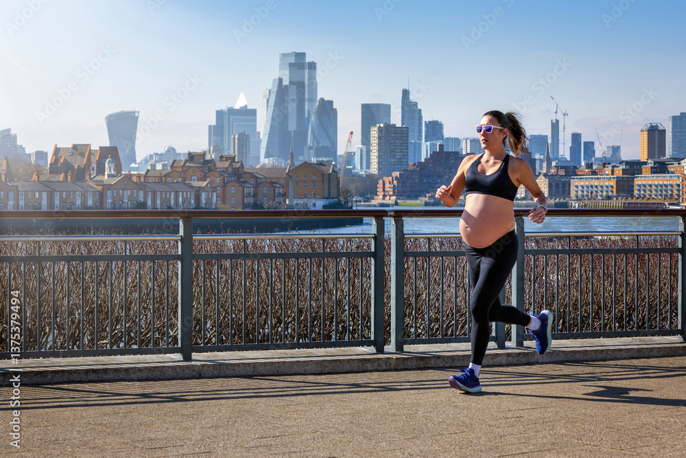 A pregnant woman in sports clothing running for exercise in front of an urban skyline during a sunny day